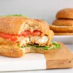 Close up on a vegan buffalo "chicken" sandwich with a bite taken off to show the middle texture from the crispy breaded tofu with the buffalo sauce dripping on the side.