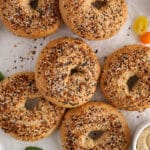Overhead view on a few vegan bagels placed on parchment paper with a bowl of hummus on the side and some fresh produce.