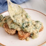 Close up on a large spoon that's serving some creamy braised white beans over 2 pieces of bread on a plate.