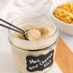 Showing is a jar filled with a vegan powdered cheese mix used to make a vegan mac and cheese. There is a bowl of vegan mac and cheese in the background.