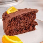 There is a slice of vegan chocolate orange cake on a plate. The cake is topped with a slice of fresh orange.