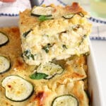 There is a large spatula holding a piece of vegan white sauce lasagna over the camera with the larger casserole int he background to show the layers.
