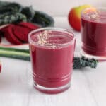 Showing are 2 glasses of beet kale smoothie with some produce in the background.