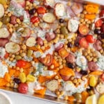 Overhead view on a vegan tray bake featuring Mediterranean ingredients such as potatoes, tomatoes, olives, chickpeas, yogurt and fresh herbs.