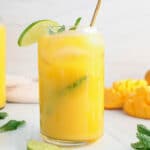 Close up on a glass of mango water filled with mint and ice cubes. There is more fresh mango in the background.