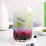 There is a tall glass of a blueberry matcha latte with more fruit and matcha powder in the background.
