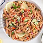 Showing is a large serving bowl filled with a vegetable and red lentil pasta salad with lemon slices.