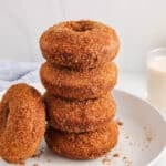 There are a few vegan apple cider donuts piled over each other on a plate with a glass of milk in the background.