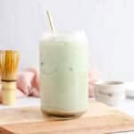 Front view on a tall glass of matcha chai latte with a matcha whisk and some maple syrup in the background.