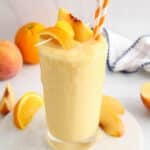 Front angle view on a tall glass of a peach pineapple smoothie topped with fresh fruits and with orange and white straws.