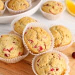 There are a few vegan cranberry orange muffins on a wooden board with more fresh cranberries around.