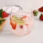 Front angle view on a small glass of strawberry kiwi water with more fresh fruit in the background.