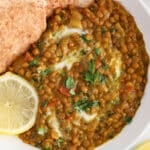Close up on a vegan brown lentil curry garnished with cilantro and served with a side of pita bread and lemon slices.