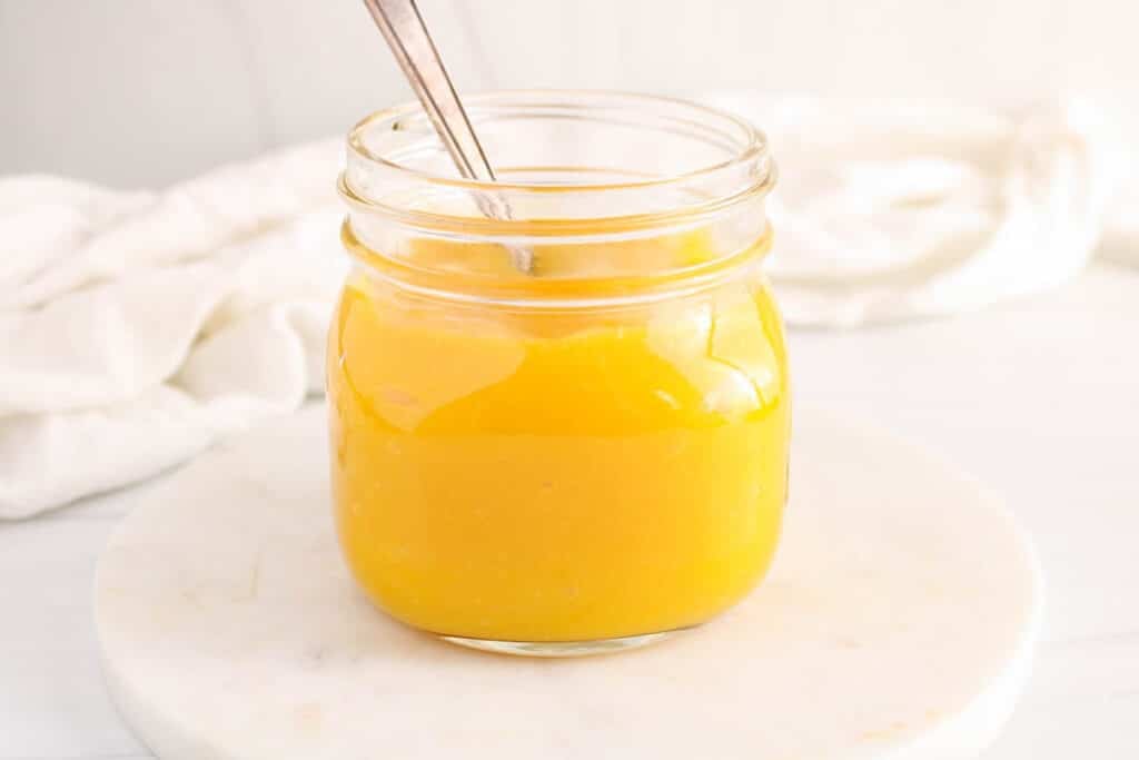 Front view on a glass jar filled with a bright yellow mango coulis with a spoon inside.