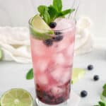 View on a tall glass of blueberry mojito mocktail with limes and fresh mint on the side.