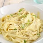 Showing a bowl of vegan fettucini Alfredo made with oat milk and garnished with green onions and ground black pepper.