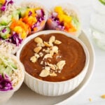 Front angle view on a small bowl of hoisin peanut sauce garnished with crushed peanuts and spring rolls on the side.