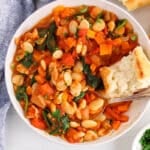 There is a spoon in a bowl of butter bean with a piece of baguette bread dipping in the tomato sauce.
