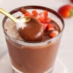 There is a spoon taking a bite of oat milk chocolate pudding from a cup.