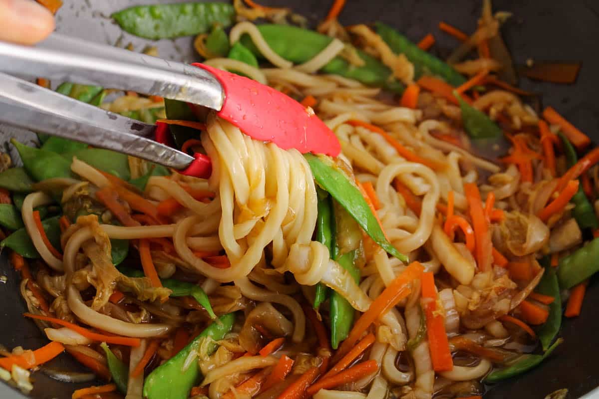 There are some tongs grabbing some udon noodles from a large pan with vegetable teriyaki.