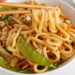 There are some chopsticks taking some noodles from a bowl of vegetable teriyaki udon noodles.