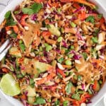 View on a large bowl of edamame crunch salad with 2 large serving spoons and a side of lime wedges.