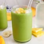 View on a tall glass of a tropical island green smoothie with more fresh fruit in the background.