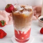Showing is a layered iced strawberry latte with ice cubes where you can see the layers of strawberry purée, milk and coffee.