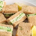 View on a large plate filled with squares of vegan cucumber sandwiches and lemon wedges.