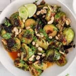 Overhead view on a bowl filled with Thai Brussels sprouts garnished with peanuts, cilantro and lime wedges.