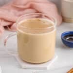 View on a mug filled with warm cinnamon milk tea with a cinnamon stick on the side and a bowl with maple syrup.