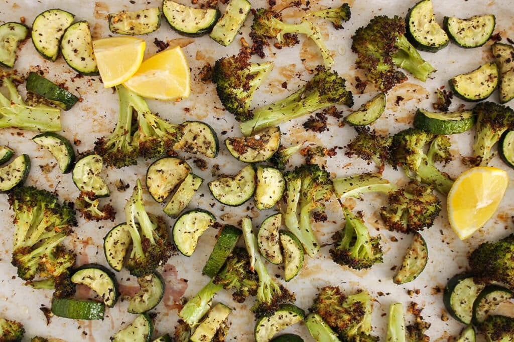 View on a large baking sheet filled with roasted zucchini and broccoli with herbs and lemon wedges.