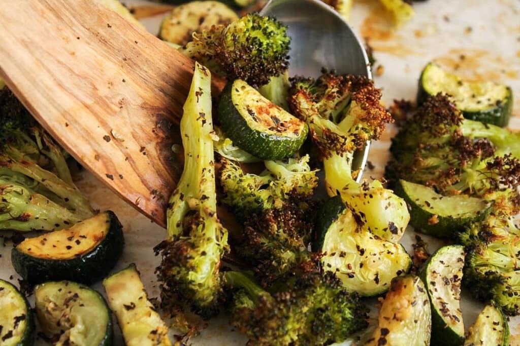 There are 2 spoons grabbing some roasted zucchini and broccoli from a sheet pan.
