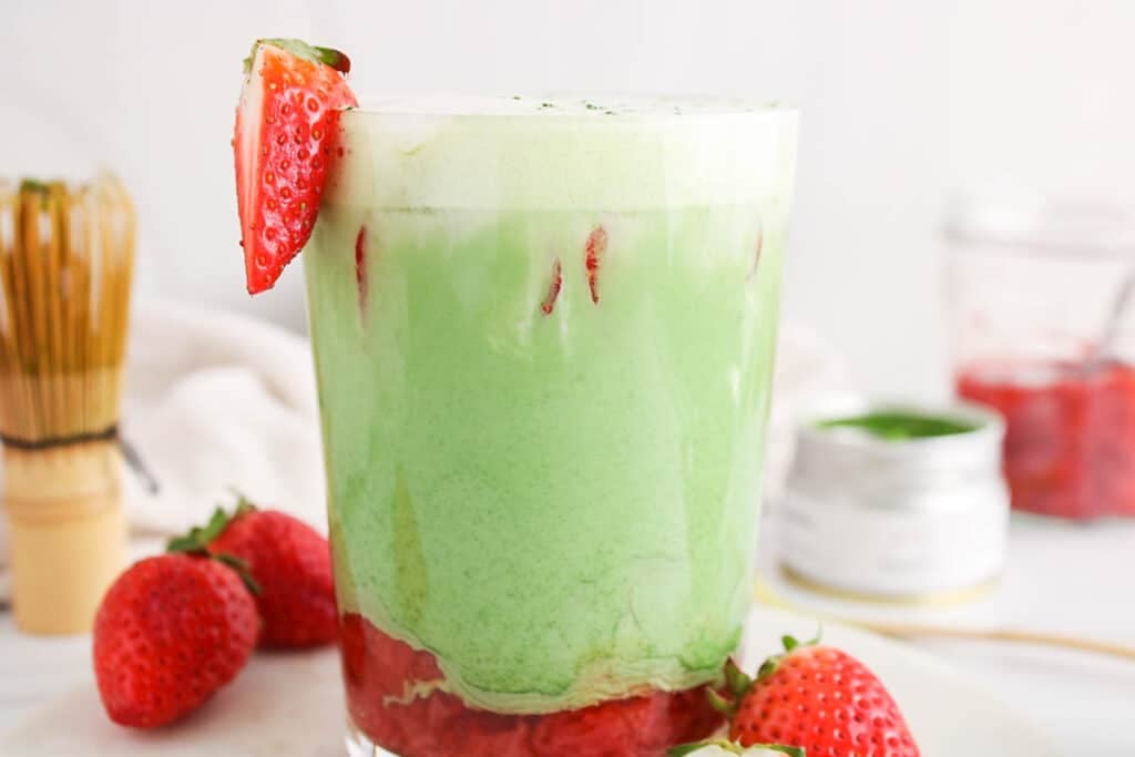 Showing is a tall glass of strawberry matcha oat latte with fresh strawberries on the side.