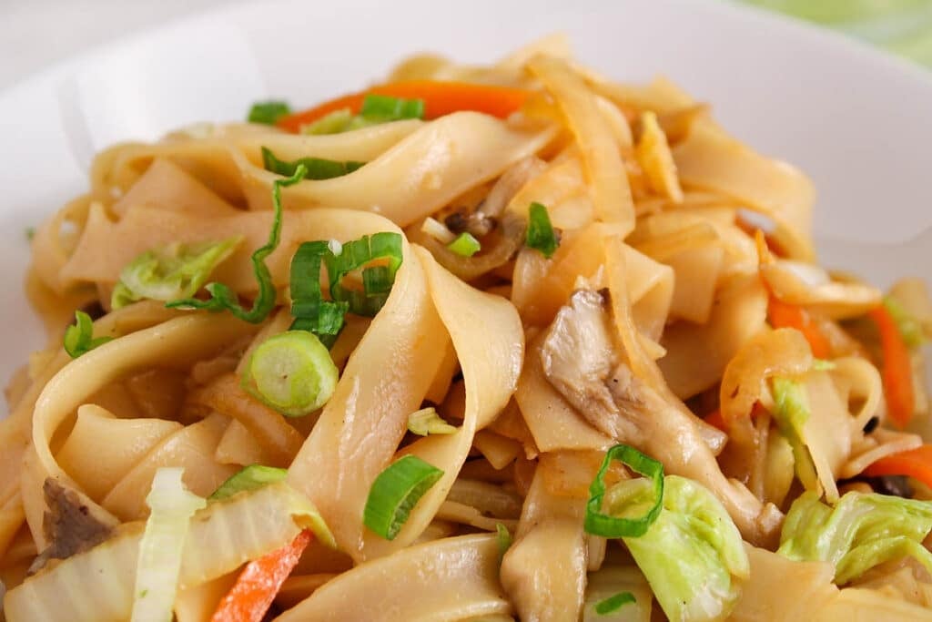 Close up on a bowl of flat rice noodles stir-fried with vegetables and soy sauce.