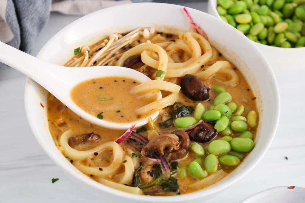 There is a spoon taking some red curry broth from a udon noodle soup bowl.