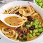 There is a spoon taking some red curry broth from a udon noodle soup bowl.