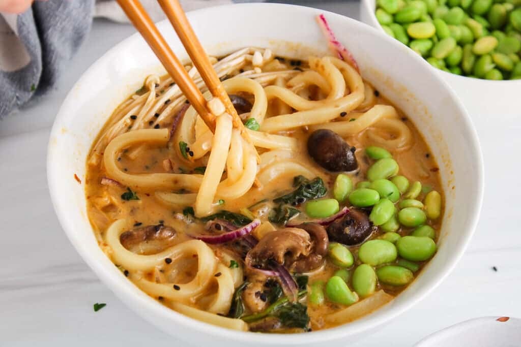There are chopsticks taking some udon noodles from a Thai red curry coconut soup.