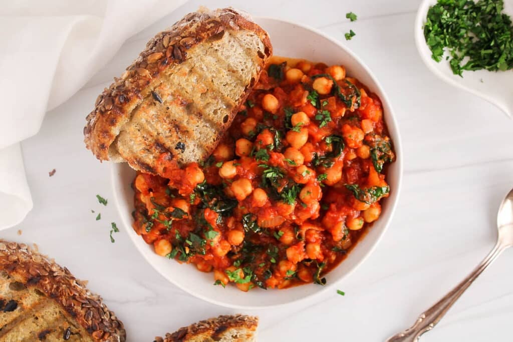 Overhead view on a bowl of chickpeas and kale in tomato sauce with a side of bread and garnished with fresh herbs.