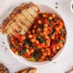Overhead view on a bowl of chickpeas and kale in tomato sauce with a side of bread and garnished with fresh herbs.