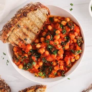 Overhead view on a bowl of chickpeas and kale in tomato sauce with a side of bread and garnished with fresh herbs.