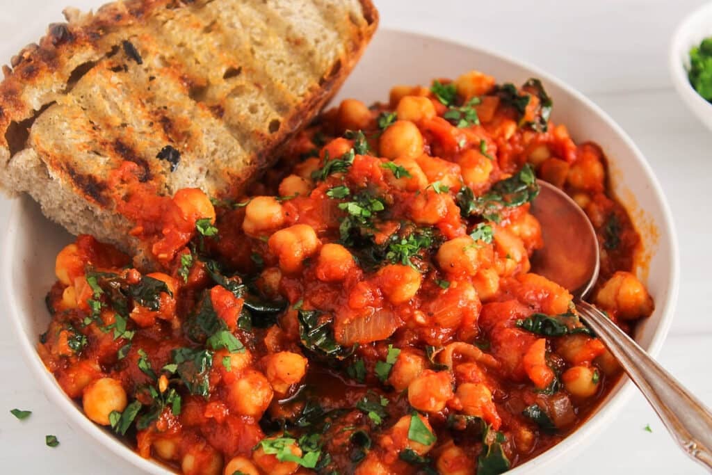 View on a bowl filled with chickpeas in tomato sauce with kale and a side of bread.