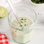 View on a jar filled with a cilantro lime tahini dressing topped with fresh herbs and with some limes in the background.