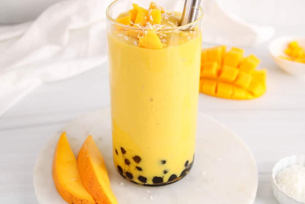 View on a tall glass of creamy tropical mango smoothie with boba and more fresh fruit on top.