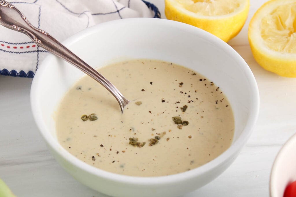 Vegan tahini cesar dressing in a bowl with a stirring spoon garnished with capers and black pepper with lemon in the background.