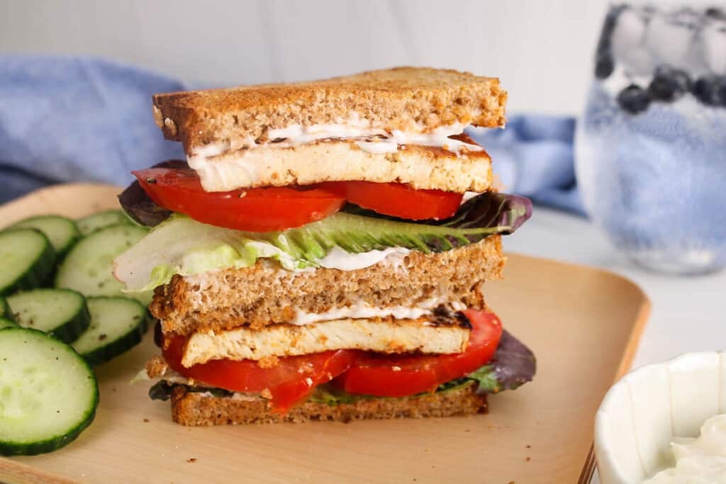 Two halves of vegan tofu BLT sandwich over each other on a plate filled with tomatoes, lettuce and mayo.