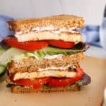 Two halves of vegan tofu BLT sandwich over each other on a plate filled with tomatoes, lettuce and mayo.