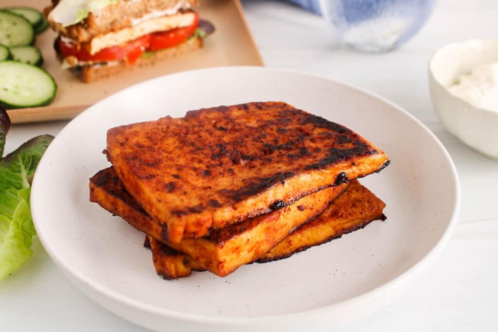 Pile of vegan tofu bacon on a white plate with a sandwich in the background.