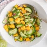 Large white bowl of cucumber mango salad in a light rice vinegar dressing with a large serving spoon.