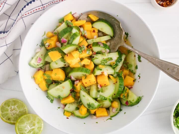 Large white bowl of cucumber mango salad in a light rice vinegar dressing with a large serving spoon.